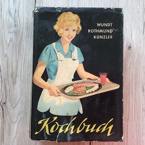 K&ouml;nnte beinhalten: Vintage Kochbuch mit schwarzem Einband. Es zeigt eine l&auml;chelnde Frau in blauem Kleid und wei&szlig;er Sch&uuml;rze, die ein Tablett mit Essen h&auml;lt. Der Titel "Kochbuch" steht in gelber Schrift, mit Autorennamen dar&uuml;ber. Das Cover hat ein Retro-Design.
