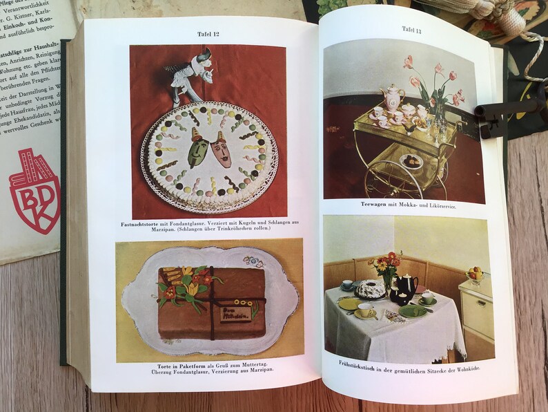 K&ouml;nnte beinhalten: Eine Seite aus einem Vintage-Kochbuch mit Illustrationen von Kuchen, einem Teeservice und einem Fr&uuml;hst&uuml;ckstisch. Die Seite enth&auml;lt deutschen Text und einen dekorativen Kuchen mit Marzipanmasken. Die Bilder sind farbenfroh und retro.