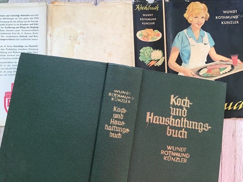 K&ouml;nnte beinhalten: Eine Sammlung alter Kochb&uuml;cher. Das Hauptbuch ist dunkelgr&uuml;n mit goldener Schrift, die "Koch und Haushaltungsbuch" und "WUNDT ROTHMUND K&Uuml;NZLER" lautet. Weitere B&uuml;cher sind sichtbar, darunter eines mit einer Frau auf dem Cover.