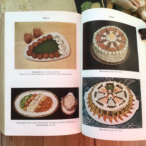 K&ouml;nnte beinhalten: Offenes Buch mit Vintage-Essensillustrationen. Eine Seite zeigt einen Teller mit Fleischb&auml;llchen, gr&uuml;nem Spinat und einer wei&szlig;en Sauce. Die andere Seite zeigt einen Kuchen mit dekorativem Design.