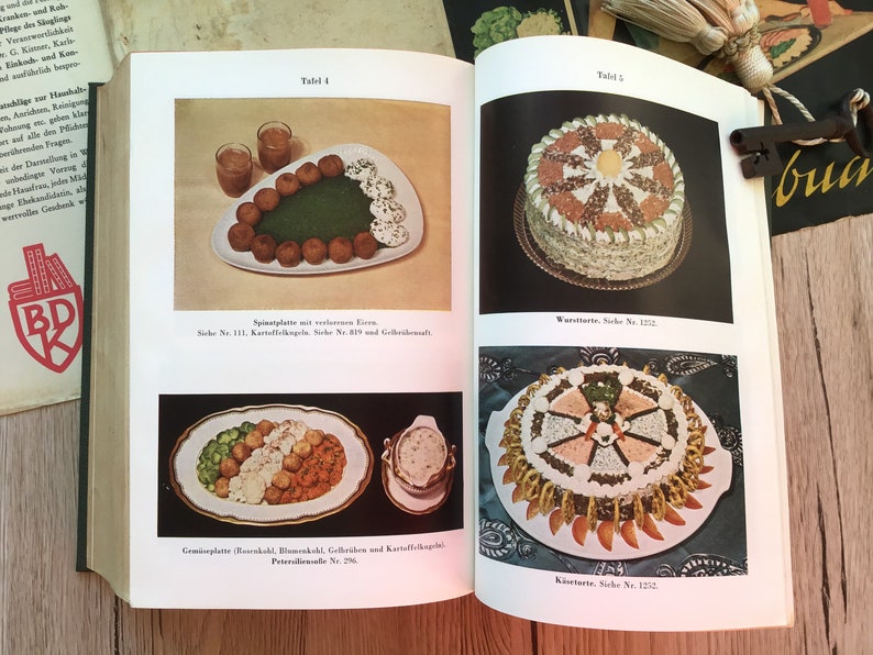 K&ouml;nnte beinhalten: Offenes Buch mit Vintage-Essensillustrationen. Eine Seite zeigt einen Teller mit Fleischb&auml;llchen, gr&uuml;nem Spinat und einer wei&szlig;en Sauce. Die andere Seite zeigt einen Kuchen mit dekorativem Design.