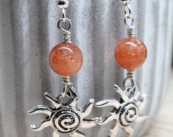 May include: A pair of dangle earrings featuring orange-red gemstone beads and silver-colored sun charms. The earrings have a hook closure and are displayed against a blurred background of greenery.