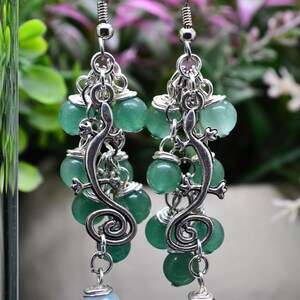 May include: A pair of silver-toned drop earrings featuring a central lizard charm with a spiral tail, accented by multiple round green beads and a single light blue bead at the bottom, all suspended from silver-toned fishhook ear wires.