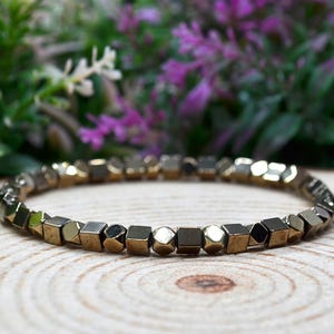 May include: A bracelet featuring geometric, metallic beads in gold and silver tones. The bracelet is circular and sits on a wooden surface. The background is blurred, showing green and purple foliage.