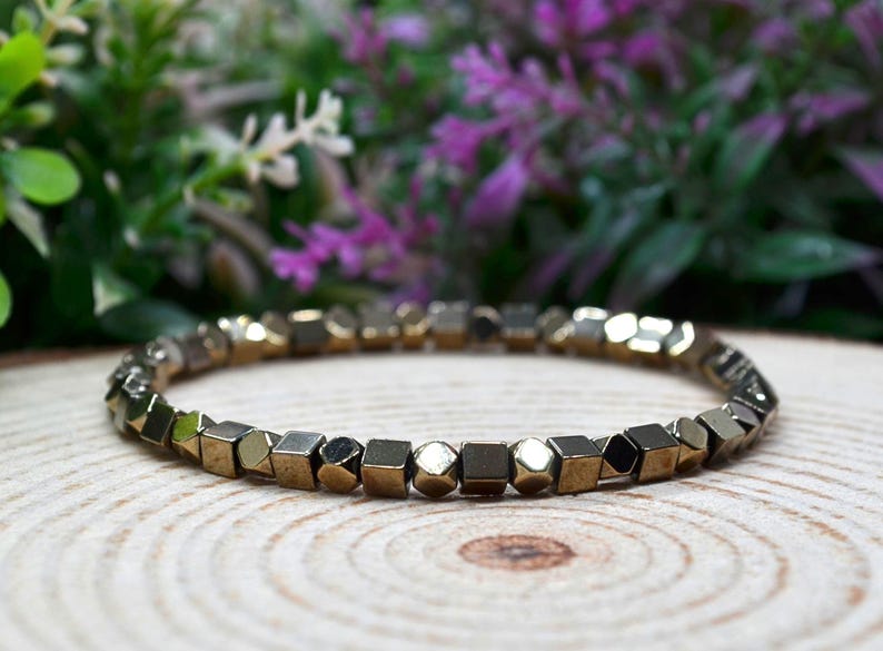 May include: A bracelet featuring geometric, metallic beads in gold and silver tones. The bracelet is circular and sits on a wooden surface. The background is blurred, showing green and purple foliage.
