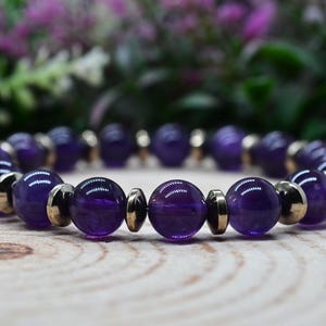 May include: A beaded bracelet featuring polished, deep purple amethyst stones and gold-colored metal accents. The bracelet is displayed on a wooden surface with a blurred floral background, showcasing the jewelry's color and design.