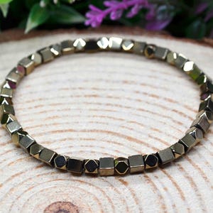 May include: A bracelet made of small, gold-coloured geometric beads. The beads have a metallic sheen and are strung together to form a circular shape. The bracelet is displayed on a wooden surface, with green and purple foliage in the background.