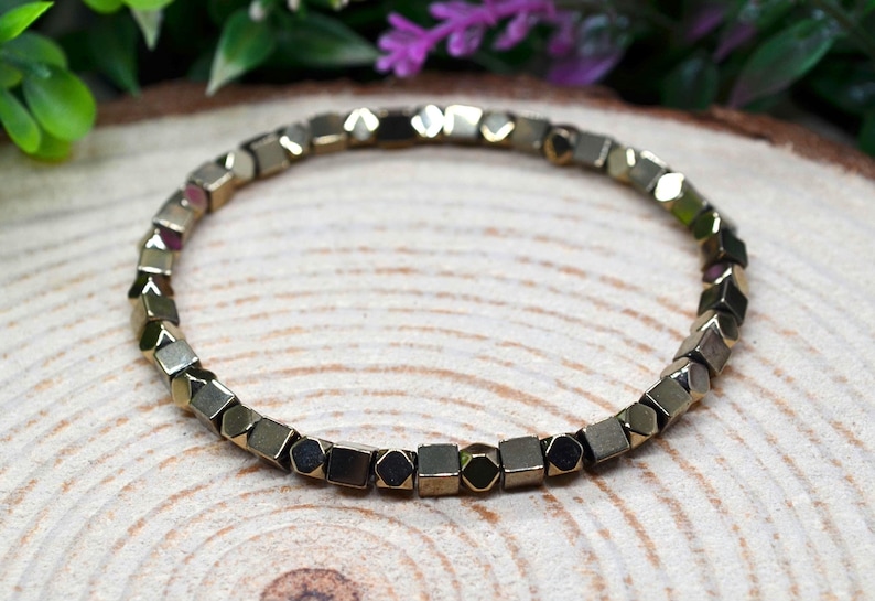 May include: A bracelet made of small, gold-coloured geometric beads. The beads have a metallic sheen and are strung together to form a circular shape. The bracelet is displayed on a wooden surface, with green and purple foliage in the background.