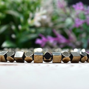May include: A gold-coloured bracelet featuring geometric beads. The beads have a metallic finish and are arranged in a row. The background is blurred, with green and purple foliage.