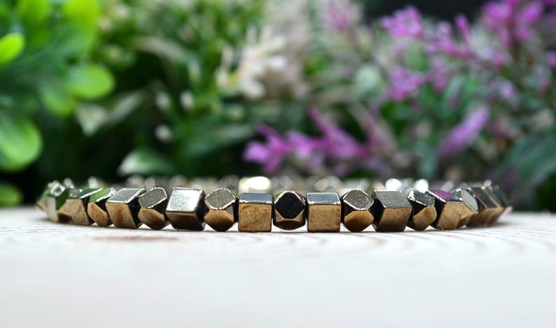 May include: A gold-coloured bracelet featuring geometric beads. The beads have a metallic finish and are arranged in a row. The background is blurred, with green and purple foliage.