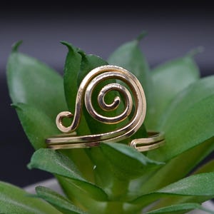 May include: A gold-toned metal ring featuring a prominent spiral design on the front. The band is open at the back, allowing for adjustment. The surface of the metal is smooth and reflective, catching the light across its curved wire form.