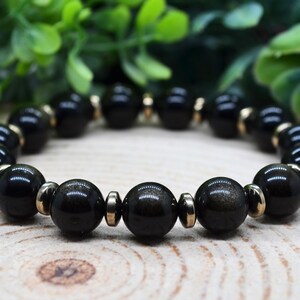May include: A beaded bracelet featuring alternating round black obsidian beads and small metallic gold-toned spacers. The polished black stones exhibit a subtle golden sheen, arranged in a circular pattern to form a continuous loop of dark beads.