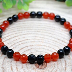 May include: A beaded bracelet featuring a repeating pattern of smooth, round, polished black and translucent orange-red gemstone beads. The beads are strung together in a circular arrangement, alternating between the two distinct colours throughout.