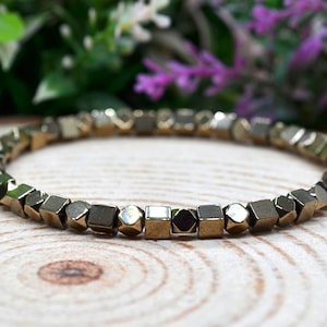 May include: A gold-coloured bracelet made of small, geometric beads. The beads have a metallic sheen and are arranged in a circular pattern. The bracelet is displayed on a wooden surface, with green and purple foliage in the background.