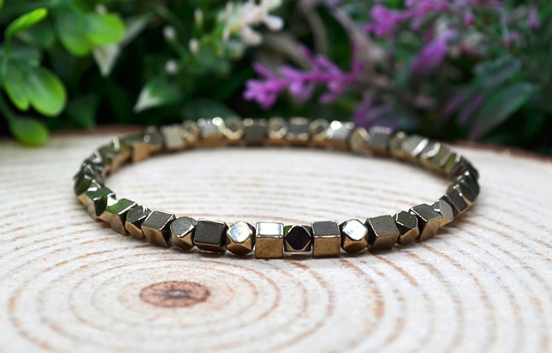 May include: A gold-coloured bracelet made of small, geometric beads. The beads have a metallic sheen and are arranged in a circular pattern. The bracelet is displayed on a wooden surface, with green and purple foliage in the background.