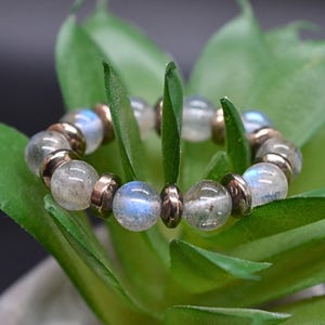 May include: A circular ring constructed from alternating round, translucent gray beads with blue iridescent flashes and small, smooth, metallic-toned disc-shaped spacers arranged in a repeating pattern around the entire circumference.