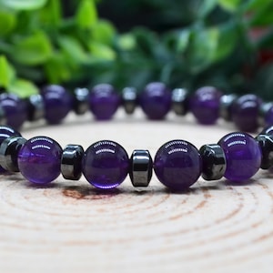 May include: A stretch bracelet featuring alternating round purple amethyst beads and smaller, metallic dark gray hematite spacer beads. The beads are arranged in a repeating pattern, creating a circular design with a polished, smooth finish.