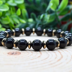 Könnte beinhalten: Ein Perlenarmband mit abwechselnd glatten, runden, polierten schwarzen Perlen und kleineren, metallisch goldfarbenen scheibenförmigen Zwischenelementen. Das Armband ist kreisförmig angeordnet und zeigt eine gleichmäßige Abfolge dunkler Kugeln.