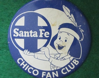 May include: A white and blue round pin with a cartoon image of a Native American holding a blue circle with the words "Santa Fe" in white. The pin has a white metal loop on top and the words "Chico Fan Club" in a blue circle around the bottom.