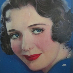 May include: A vintage magazine cover featuring a woman with dark hair and a bright red lipstick. The magazine is called "Photoplay" and the cover features the text "The "New Deal" in Girls" and the name "Ruby Keeler".