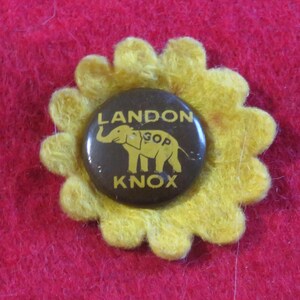 May include: A brown button with a yellow felt flower around it. The button has a picture of an elephant with the text "LANDON GOP KNOX" on it.
