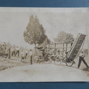 May include: A black and white vintage photograph of a steam-powered threshing machine in a field. The machine is being operated by several people, and there are horses in the background.