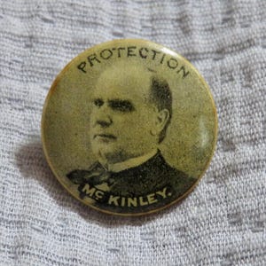 May include: A vintage round button with a portrait of a man in a suit. The button has the word "PROTECTION" arched across the top and "MS KINLEY" at the bottom. The button is cream-colored.