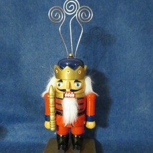 May include: A wooden nutcracker figurine with a red and blue uniform, a gold crown, and a white beard. The nutcracker is holding a gold-colored weapon and is standing on a gold base.