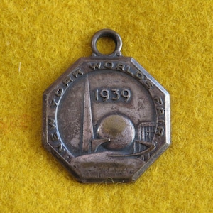 May include: A silver-toned metal pendant with an octagonal shape. The pendant features an image of the Trylon and Perisphere, the iconic symbols of the 1939 New York World's Fair. The text "NEW YORK WORLD'S FAIR 1939" is inscribed around the edge of the pendant.
