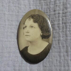May include: Oval-shaped, sepia-toned portrait of a woman. The woman has short, curly hair and is wearing a dark-colored dress. The portrait is set against a light gray textured background.