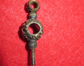 May include: A gold-toned watch key with intricate details and a round, decorative head. The key has a long, thin shaft.