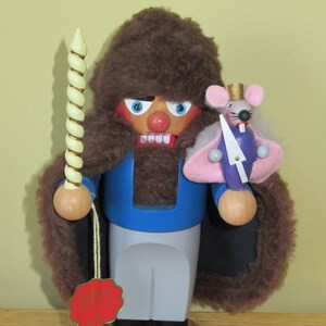 May include: A wooden nutcracker with a brown fur coat, blue jacket, and gray pants. He is holding a white and yellow candy cane in one hand and a small, pink and blue mouse nutcracker in the other. The nutcracker is standing on a wooden base.