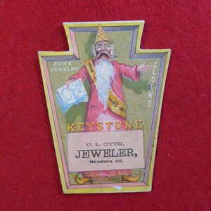 May include: A vintage advertisement for C.L. Otto, a jeweler in Mendota, Illinois. The ad features a cartoon illustration of a man with a long beard and a pointed hat, holding a watch and a pair of scissors. The text reads "Fine Jewelry, Clocks, Keystone, C.L. Otto, Jeweler, Mendota, Ill., Watch Cases Repairing."