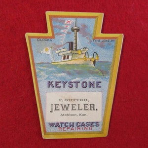 May include: Vintage Keystone Jeweler advertisement card. The card features a ship illustration with the text "KEYSTONE" and "F. Sutter, Jeweler, Atchison, Kan." The card also mentions "CLOCKS", "FINE JEWELRY", and "WATCH CASES REPAIRING". The card is on a red background.