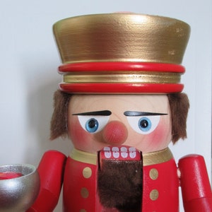 May include: A wooden nutcracker figurine with a red and gold uniform, holding a silver goblet. The nutcracker has a painted face with blue eyes, a red nose, and a brown beard.