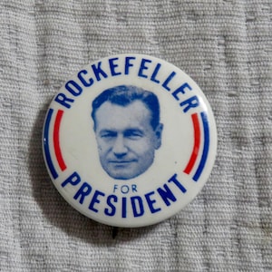 May include: A white campaign button with the text "ROCKEFELLER FOR PRESIDENT" in blue lettering. The button features a portrait of a man in a blue and red circular design.