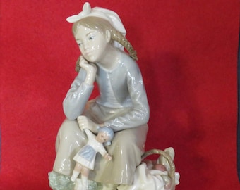 May include: A porcelain figurine of a seated girl with braided hair, wearing a light blue top and a white bonnet. She holds a doll and sits near a basket. The figurine has a serene expression and is set on a base with floral details.