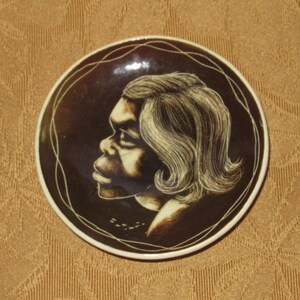 May include: A brown ceramic plate with a white design of a person's face with a side profile. The person has dark hair and is wearing a white shirt.