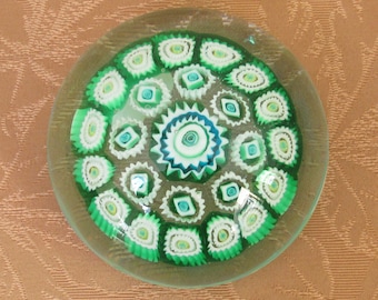 May include: A clear glass paperweight with a detailed, colorful design. The central design features a blue and white starburst, surrounded by green and white floral patterns. The paperweight is round and sits on a neutral background.