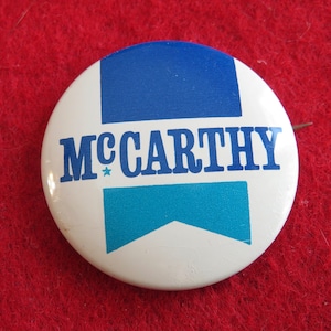 May include: A round, white button with the name "McCarthy" in blue lettering. The button has a blue and teal design, with a star next to the name. The button is pinned to a red surface.