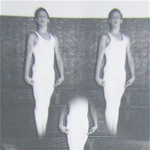May include: Black and white photo of three people wearing white tank tops. The people are standing in a row, with the person in the middle blurred out. The people are standing in front of a brick wall.