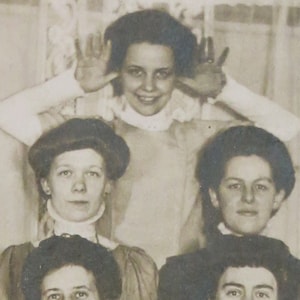 May include: Black and white vintage photograph of four women. The woman in the center is covering her ears with her hands and smiling. The other three women are looking at the camera.