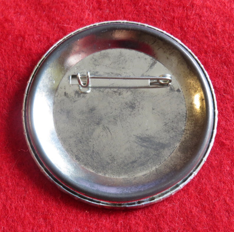 May include: The back of a silver metal button with a pin attached. The button is round and has a smooth surface.