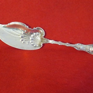 May include: Silver-colored cake server with an ornate handle and a wide, curved blade. The blade features a decorative floral design. The handle is detailed with intricate patterns. The server is likely vintage or antique.
