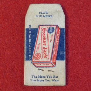 Może przedstawiać: Winietkowe pudełko Cracker Jack z nadrukiem "Blow for More" i "The Cracker Jack Co." na przodzie. Pudełko jest czerwone, białe i niebieskie z niebiesko-białym wzorem w paski. Na dole pudełka znajduje się napis "The More You Eat The More You Want".