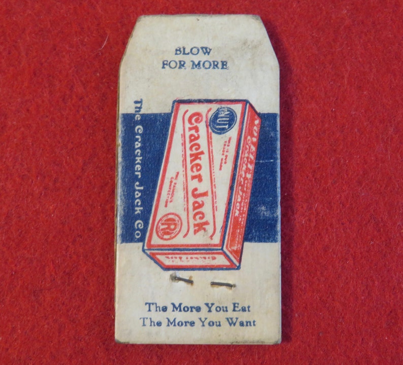 Może przedstawiać: Winietkowe pudełko Cracker Jack z nadrukiem "Blow for More" i "The Cracker Jack Co." na przodzie. Pudełko jest czerwone, białe i niebieskie z niebiesko-białym wzorem w paski. Na dole pudełka znajduje się napis "The More You Eat The More You Want".