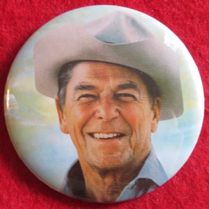 May include: A close-up of a smiling man wearing a light gray cowboy hat. He is wearing a light blue shirt and has a white background.