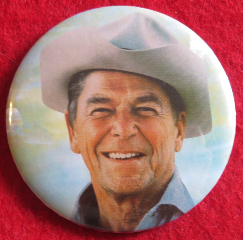 May include: A close-up of a smiling man wearing a light gray cowboy hat. He is wearing a light blue shirt and has a white background.