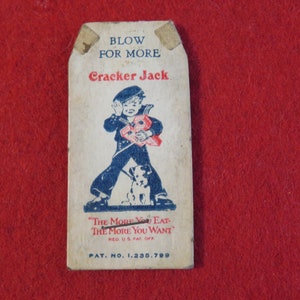 Może przedstawiać: Wierzchnia część vintage'owego pudełka Cracker Jack z niebiesko-białą ilustracją chłopca w marynarskim stroju trzymającego czerwono-białe pudełko. Tekst na pudełku brzmi "Blow for More Cracker Jack" i "The More You Eat The More You Want".