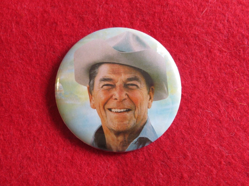 May include: A round button with a portrait of a man wearing a cowboy hat. The man is smiling and has white hair. The background is a light blue sky with clouds.